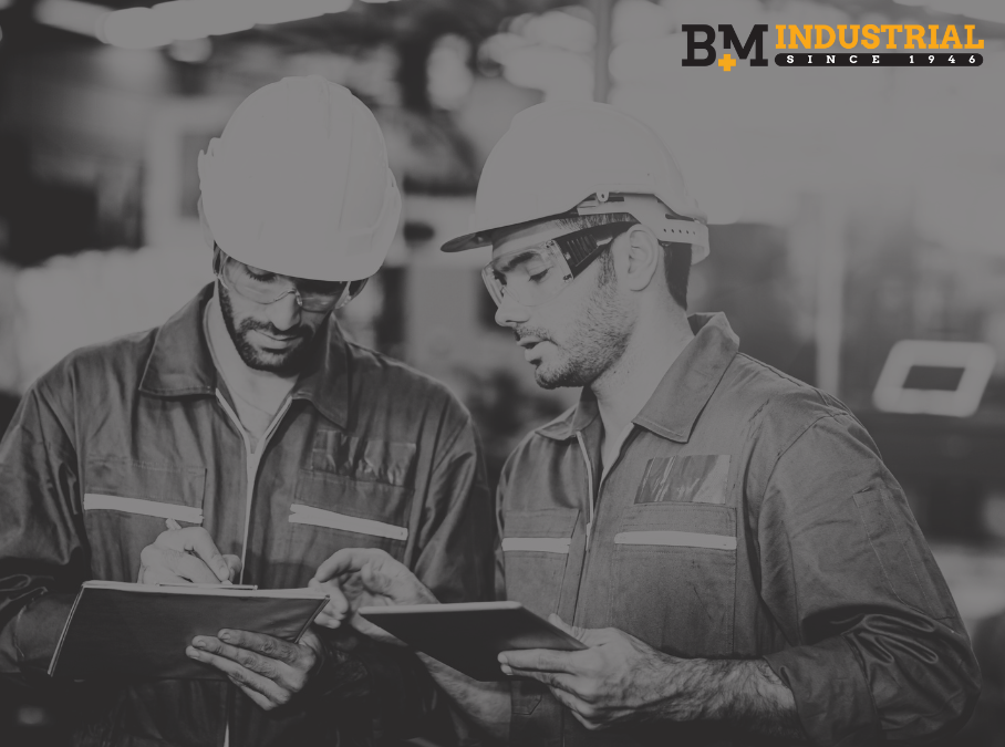 Black and white image of two industrial workers wearing hard hats and safety glasses, reviewing notes on a clipboard and tablet, with the B+M Industrial logo in the corner.